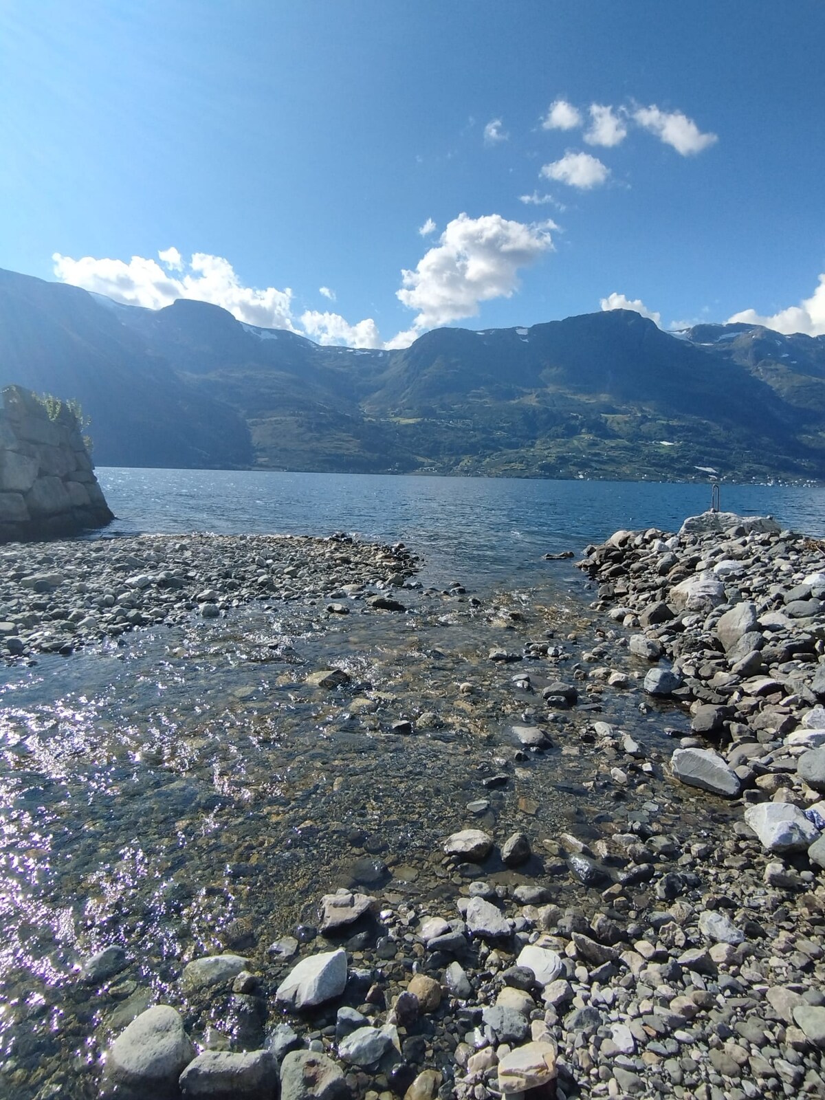 A serene lakeside scene features a rocky shoreline leading into clear waters. Gentle ripples reflect the bright blue sky, while distant hills and mountains provide a scenic backdrop. Various sizes of smooth stones create a natural pathway along the water's edge.