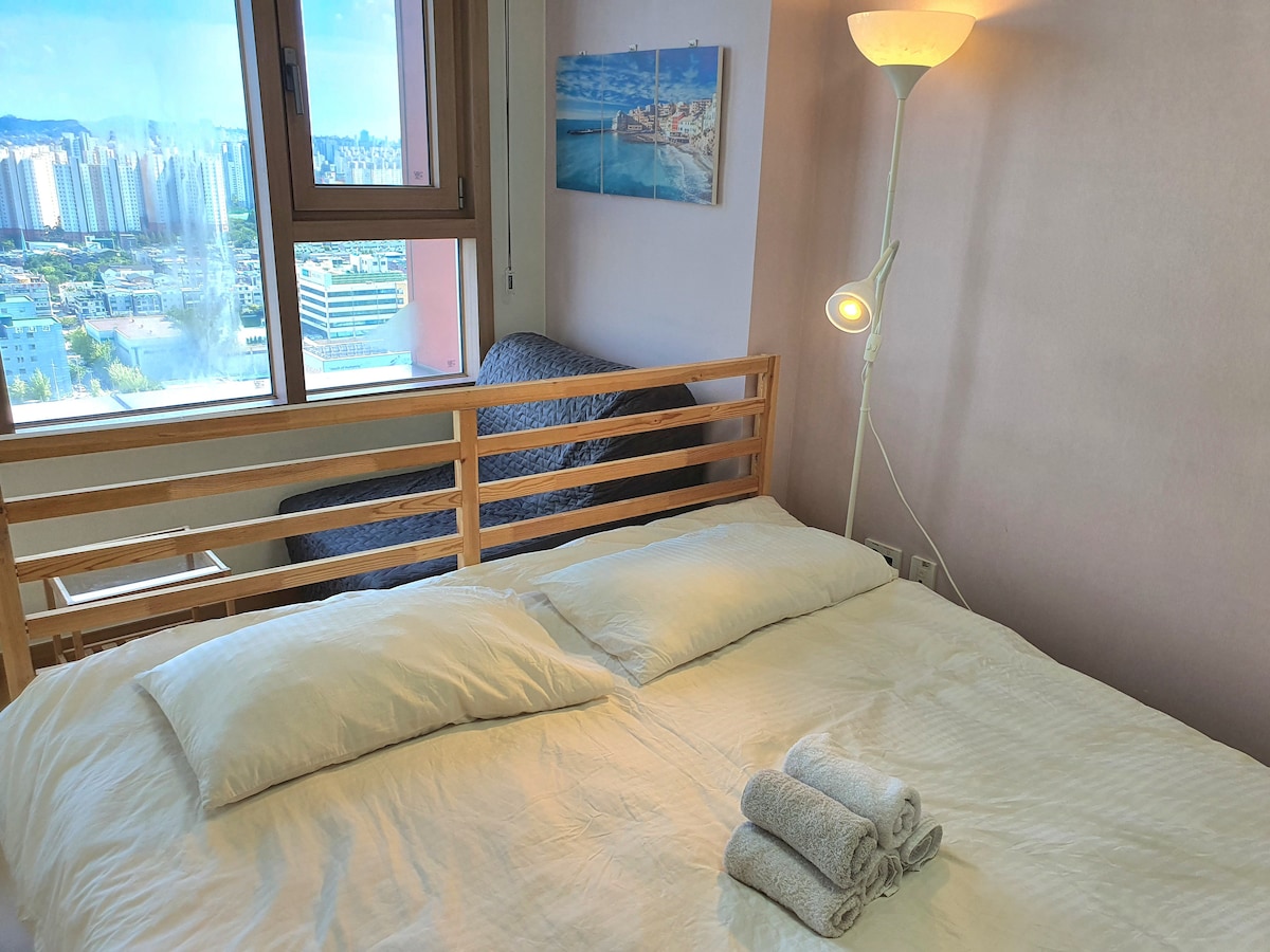 A well-lit bedroom features a spacious bed with neatly arranged towels. A modern floor lamp illuminates the room, while a nearby window offers a view of the city skyline. A cozy seating area is situated adjacent to the window, enhancing the room’s welcoming feel.