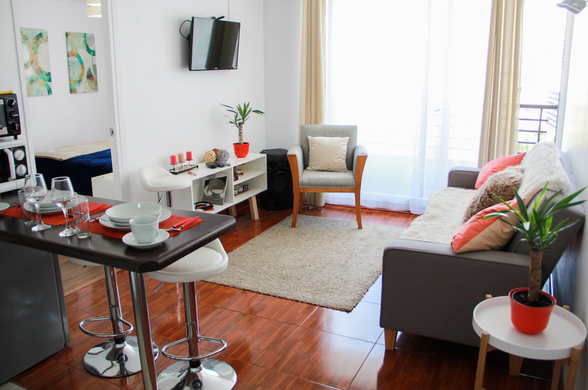 A bright living area features a comfortable gray couch and a wooden table set for meals. Natural light fills the space through large windows, accompanied by decorative plants. A small flat-screen TV is mounted on the wall, while a cozy armchair adds additional seating.