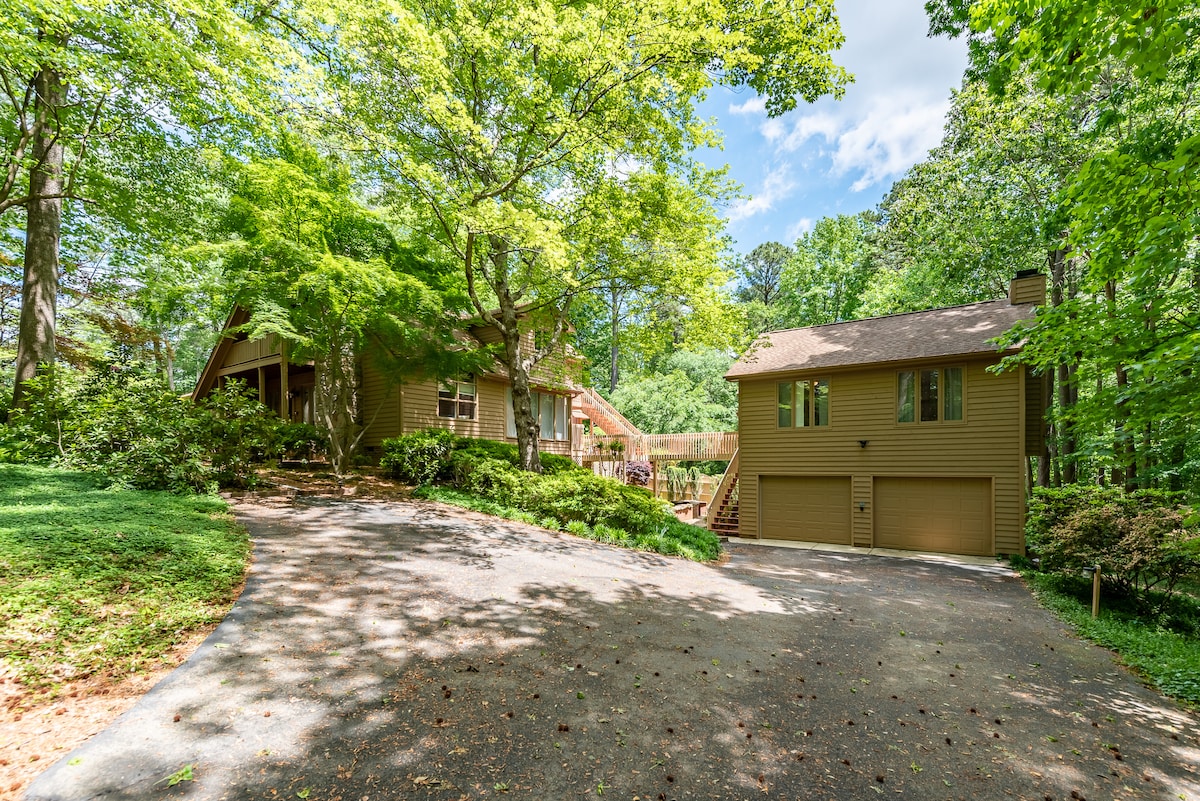 A serene exterior view of a private home surrounded by lush greenery. The driveway leads to a two-car garage attached to a detached guest suite, while stairs ascend to a spacious deck overlooking the natural landscape.