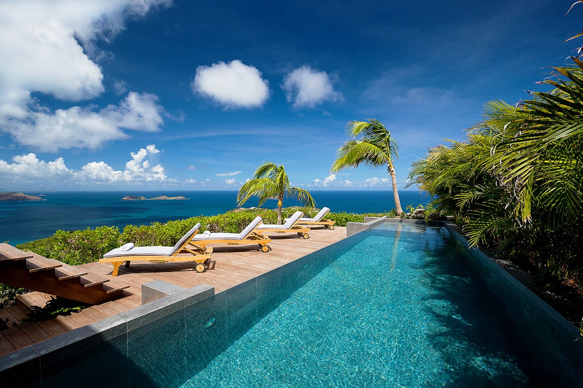A serene swimming pool is nestled among tropical foliage, showcasing several padded sun loungers arranged along a wooden deck. The expansive ocean view is complemented by a clear blue sky dotted with fluffy white clouds.