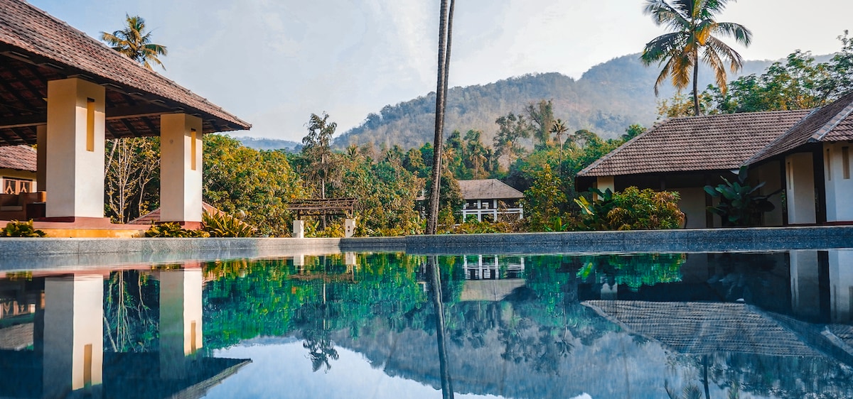 A tranquil pool reflects the surrounding greenery and mountains, with palm trees framing the view. The bungalow structures are visible in the background, blending harmoniously with the natural landscape.