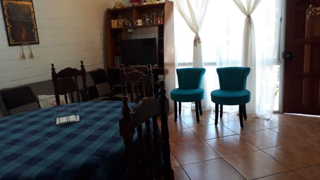 A dining area is featured, showcasing a large, checkered table surrounded by dark wooden chairs. Two vibrant blue chairs are positioned near a window, with sheer curtains allowing natural light to fill the space. A cozy sofa is seen in the background.