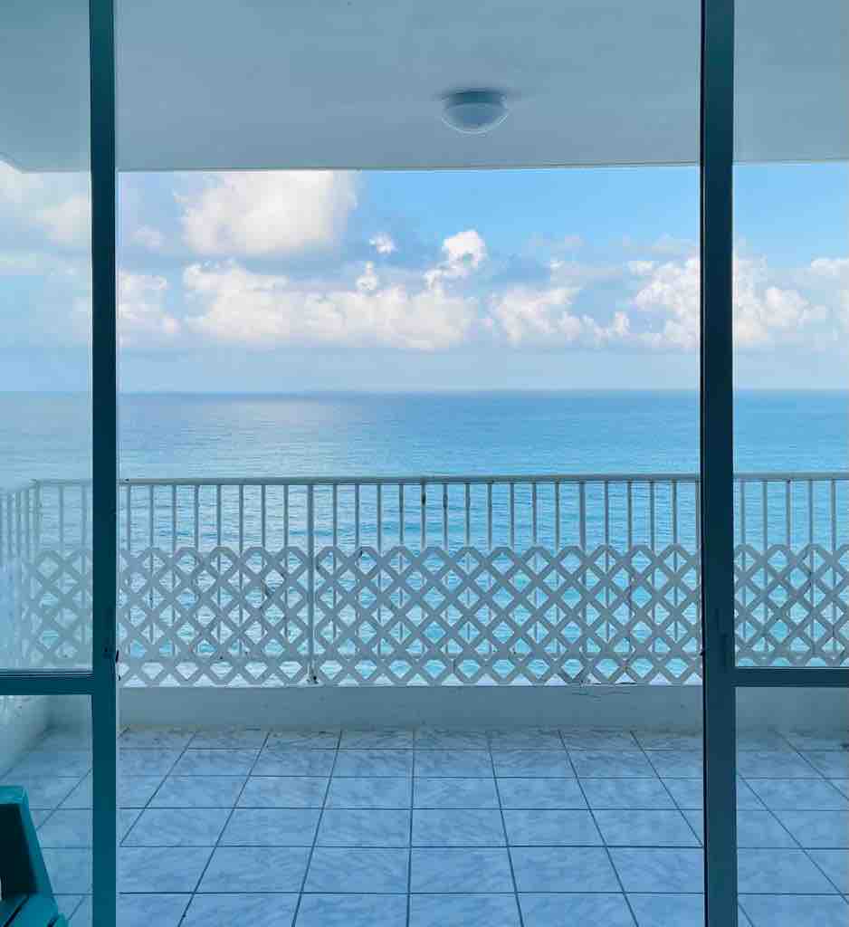A balcony is visible, featuring a white lattice railing that frames a serene view of the ocean. The sky is softly illuminated with clouds, and the floor is tiled, creating a spacious area for relaxation.