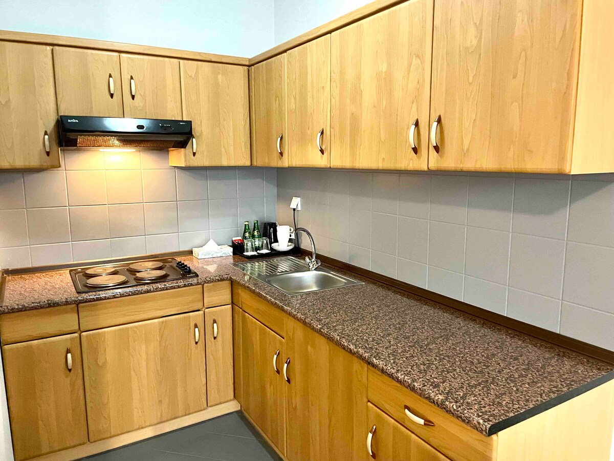 A kitchen area is presented with wooden cabinetry and a granite countertop. A stove, oven, and sink are integrated into the counter space. Bottled drinks and kitchen utensils are visible on the countertop. Lighting above the stove highlights the workspace.