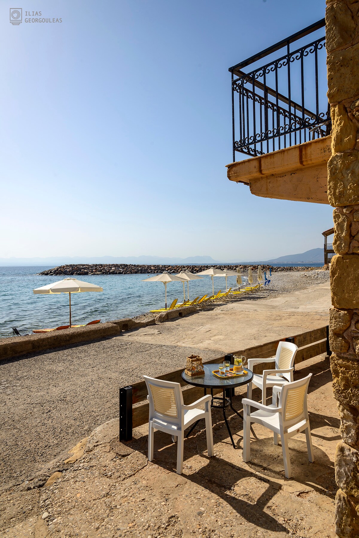 A charming outdoor seating area is set up near the beach, featuring a small table with two chairs. Sun loungers and umbrellas are visible along the sandy shore, with serene waves lapping nearby. A glimpse of the tranquil sea can be seen in the background.