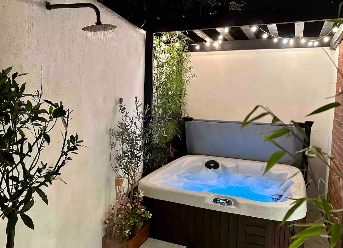 A private hot tub is nestled within a Mediterranean styled courtyard, surrounded by lush greenery and decorative plants. Soft lighting from overhead fairy lights enhances the serene atmosphere, while steam rises gently from the bubbling water, indicating a warm and inviting temperature.