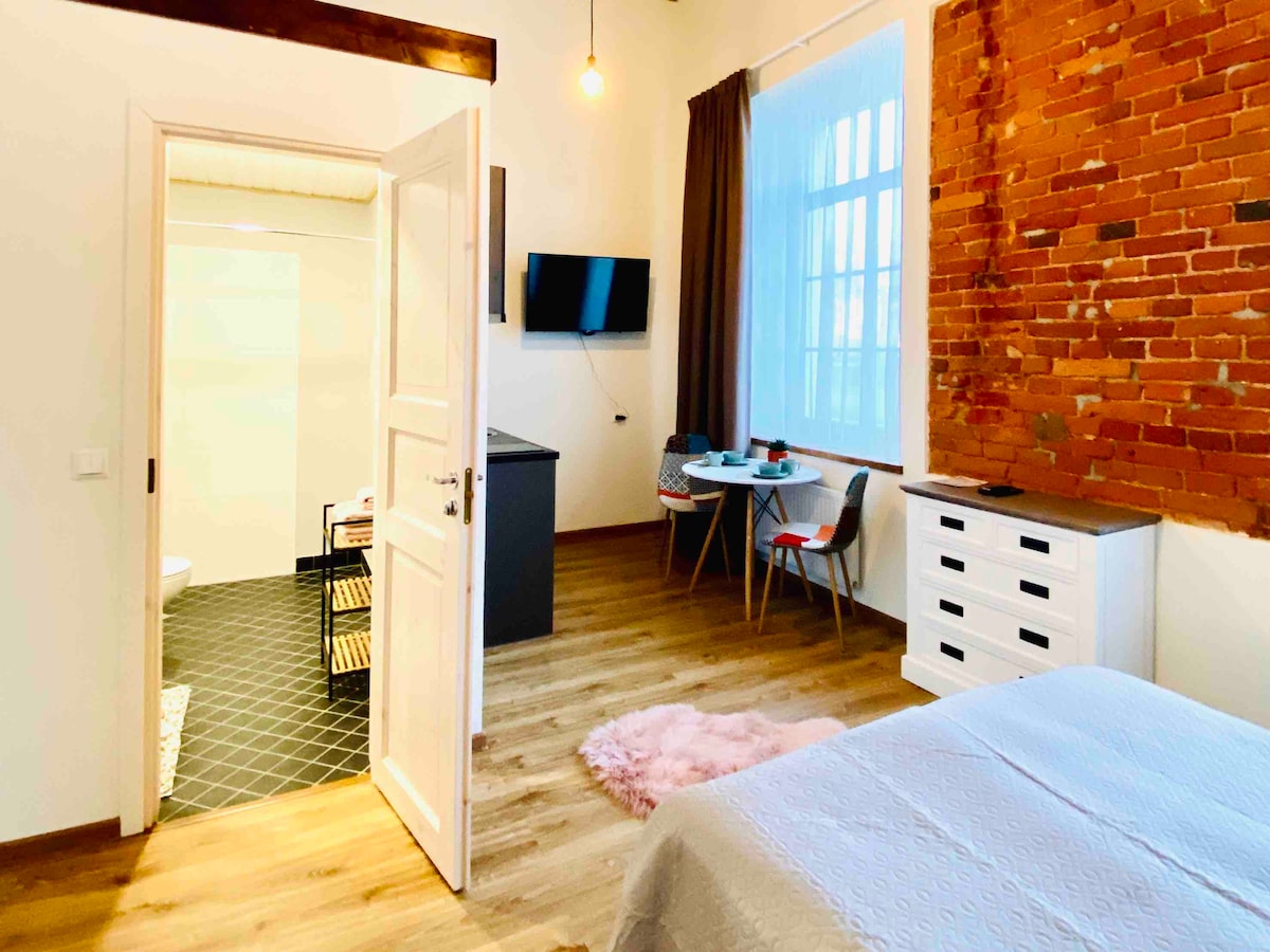 The studio apartment features a cozy sleeping area with a light-toned bedspread and a wooden floor. Exposed brick walls add character, complemented by a small dining table and chairs. A television is mounted on the wall, and natural light floods through large windows.