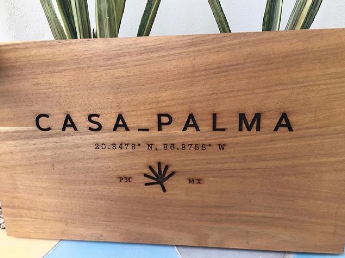 A wooden sign displaying the name 'CASA PALMA' is shown. The sign features coordinates of the location, a palm tree graphic, and the initials 'PM' and 'MX' for Puerto Morelos, Mexico. The natural wood texture adds a rustic charm to the entrance.