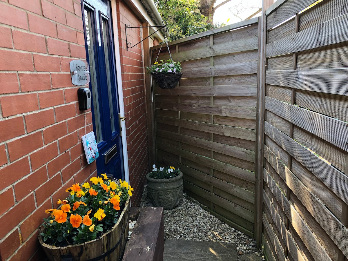 A welcoming entrance is framed by wooden fencing and brick walls, featuring a decorative blue door. Pansies in a barrel add a splash of color, while hanging flower baskets enhance the space. Gravel pathways lead through a cozy outdoor area, inviting guests to explore.