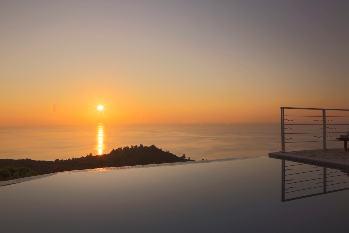 A serene sunset is captured over the Ionian Sea, with warm hues of orange and yellow reflecting off the calm water. The stillness of the private pool is highlighted by its edge merging with the ocean view, creating a tranquil atmosphere.
