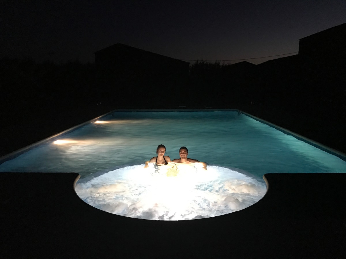 A tranquil nighttime scene featuring a well-lit pool with an integrated jacuzzi. Soft lighting reflects off the water's surface, creating a soothing atmosphere. Two individuals are seated in the jacuzzi area, surrounded by a serene outdoor environment.