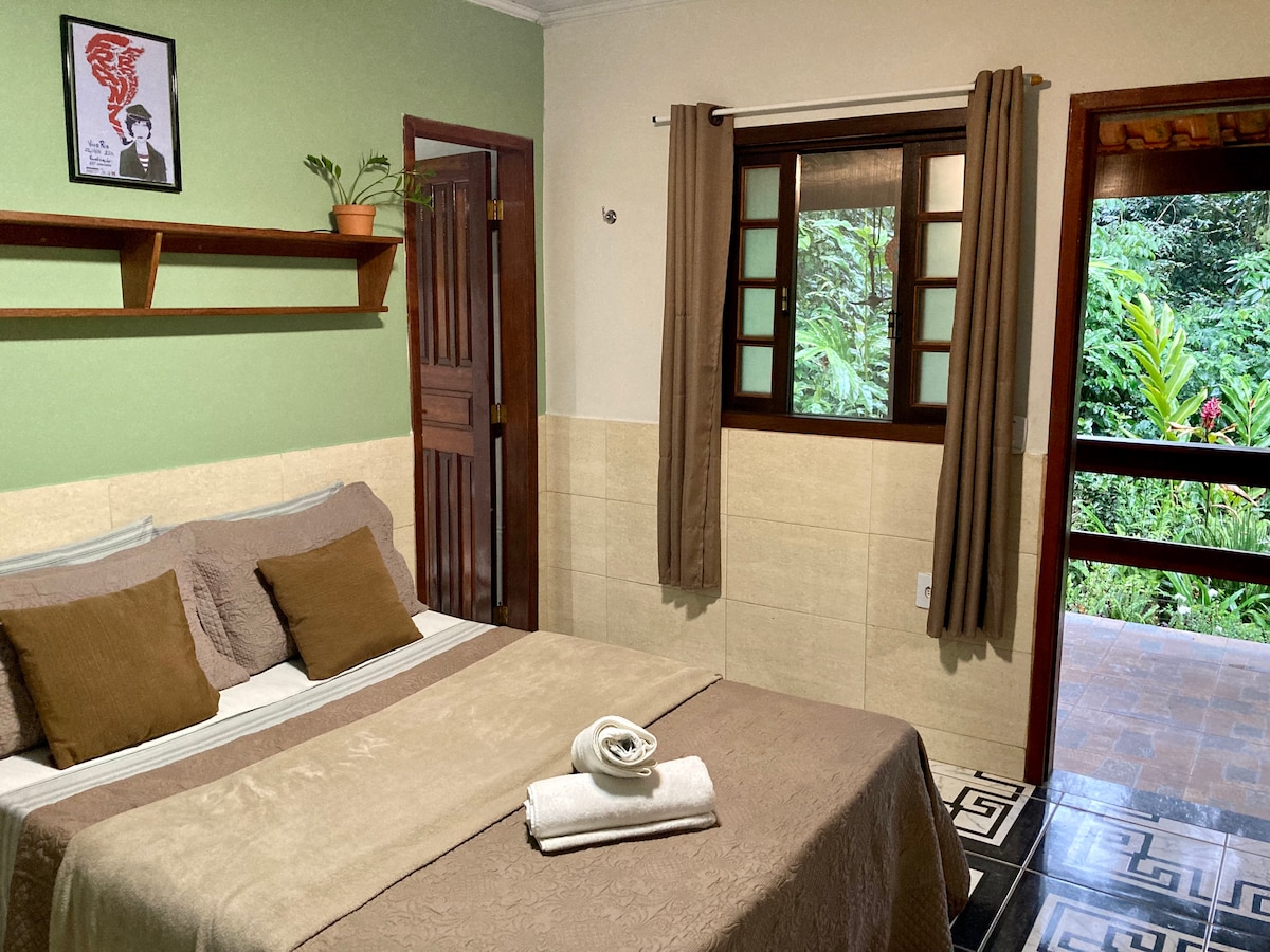 A well-lit room features a spacious bed adorned with neutral linens and decorative pillows. Wooden shelves hold a small plant and framed art. A window reveals a view of the surrounding greenery, while the inviting decor emphasizes a serene atmosphere.