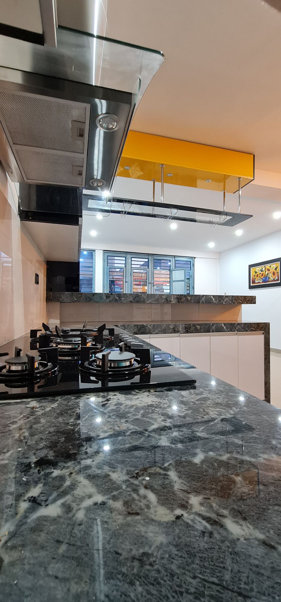 The modern kitchen features a dark marble countertop with a gas stove and sleek cabinetry. Ample overhead lighting illuminates the space, highlighting the contemporary design. A glass barrier separates the kitchen from the living area, allowing for an open feel while maintaining distinct zones.