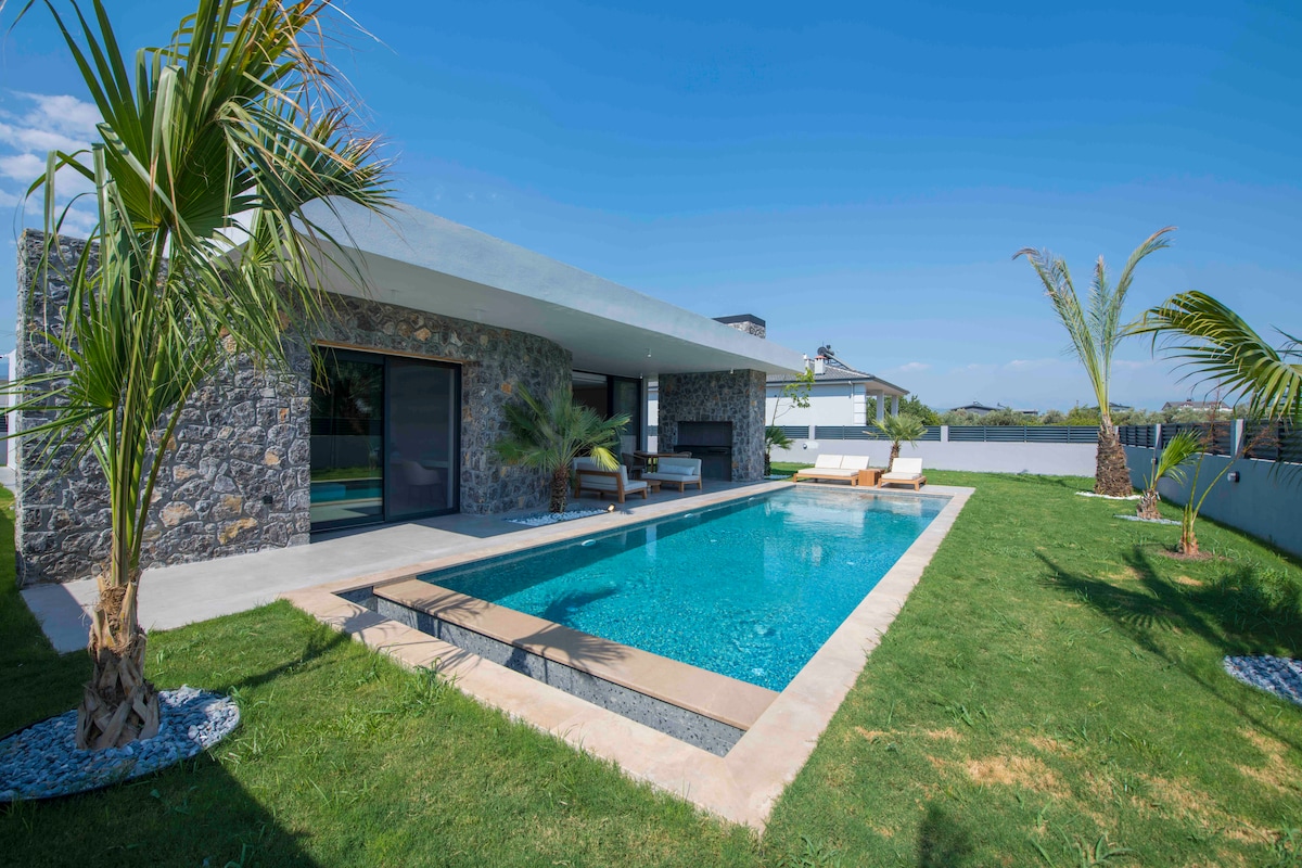 A modern villa exterior is framed by lush greenery, featuring a serene pool with crystal-clear water. Sun loungers are positioned beside the pool, inviting relaxation. The villa's stone facade complements the natural surroundings under a clear blue sky.