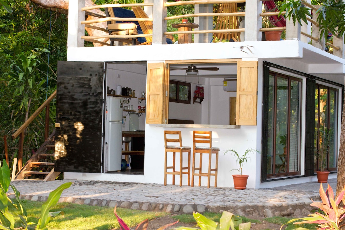 The image captures the exterior of the two-story house, featuring an open layout. A bar area with wooden stools is positioned beneath large, sliding glass doors. Tropical foliage surrounds the patio, with a staircase leading to the upper level, creating a seamless transition to nature.