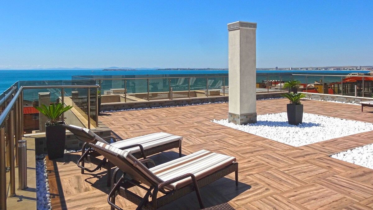 The rooftop terrace features sun loungers positioned for relaxation, with a clear view of the sea. A decorative planter and a stone structure add visual interest to the space, surrounded by a modern deck design. The open sky enhances the feeling of spaciousness.