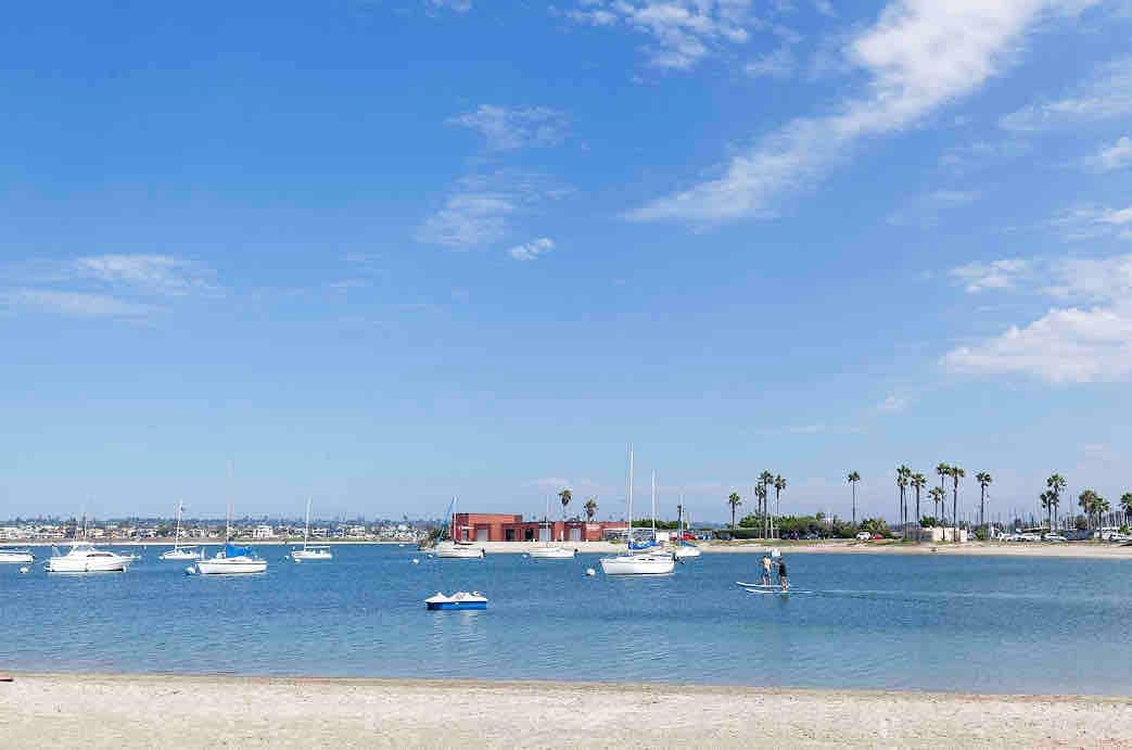 A serene waterfront scene features calm blue waters dotted with multiple sailboats. Palm trees line the shore, creating a relaxed coastal atmosphere. In the background, a distinctive red building is visible, contrasting against the sky, which is filled with light clouds.
