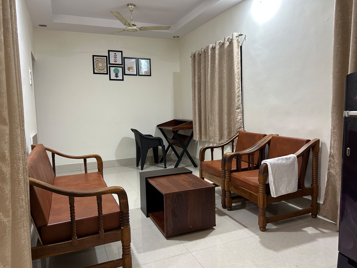 A living area is furnished with two wooden chairs and a matching sofa, complemented by a wooden coffee table at the center. A desk and black chair are positioned against the wall, and framed artworks hang above. Curtains frame a window, allowing natural light to enter.