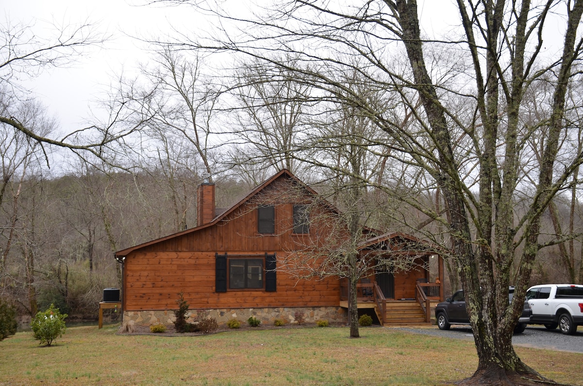 The cabin's exterior is showcased, featuring natural wood siding and large windows. Leafless trees frame the property, contributing to the serene environment. A gravel driveway leads to the entrance, providing access to the spacious yard surrounding the home.