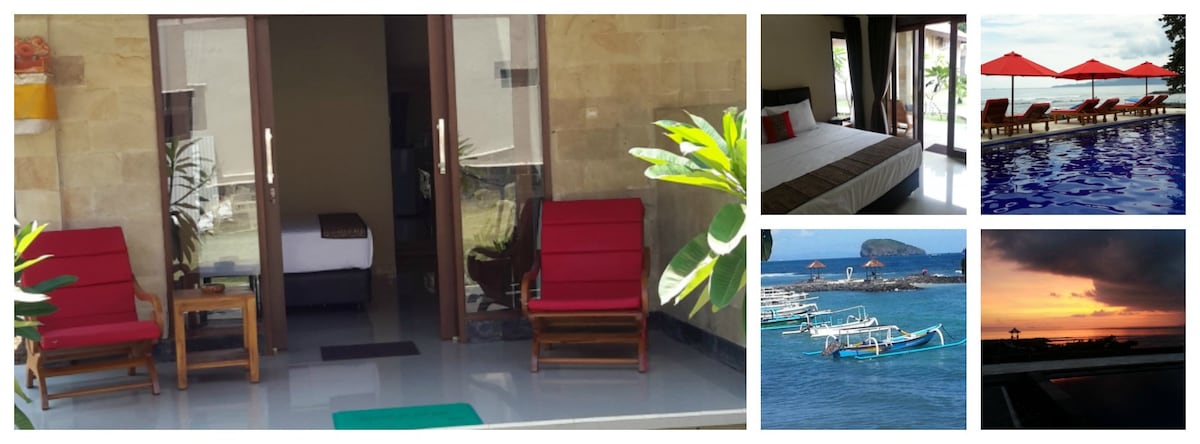 A welcoming entrance features two red lounge chairs positioned outside sliding glass doors. The dark wooden table between them complements the sleek floor. The interior room is visible, showcasing a comfortable bed with neutral bedding, while tropical plants add a touch of greenery.