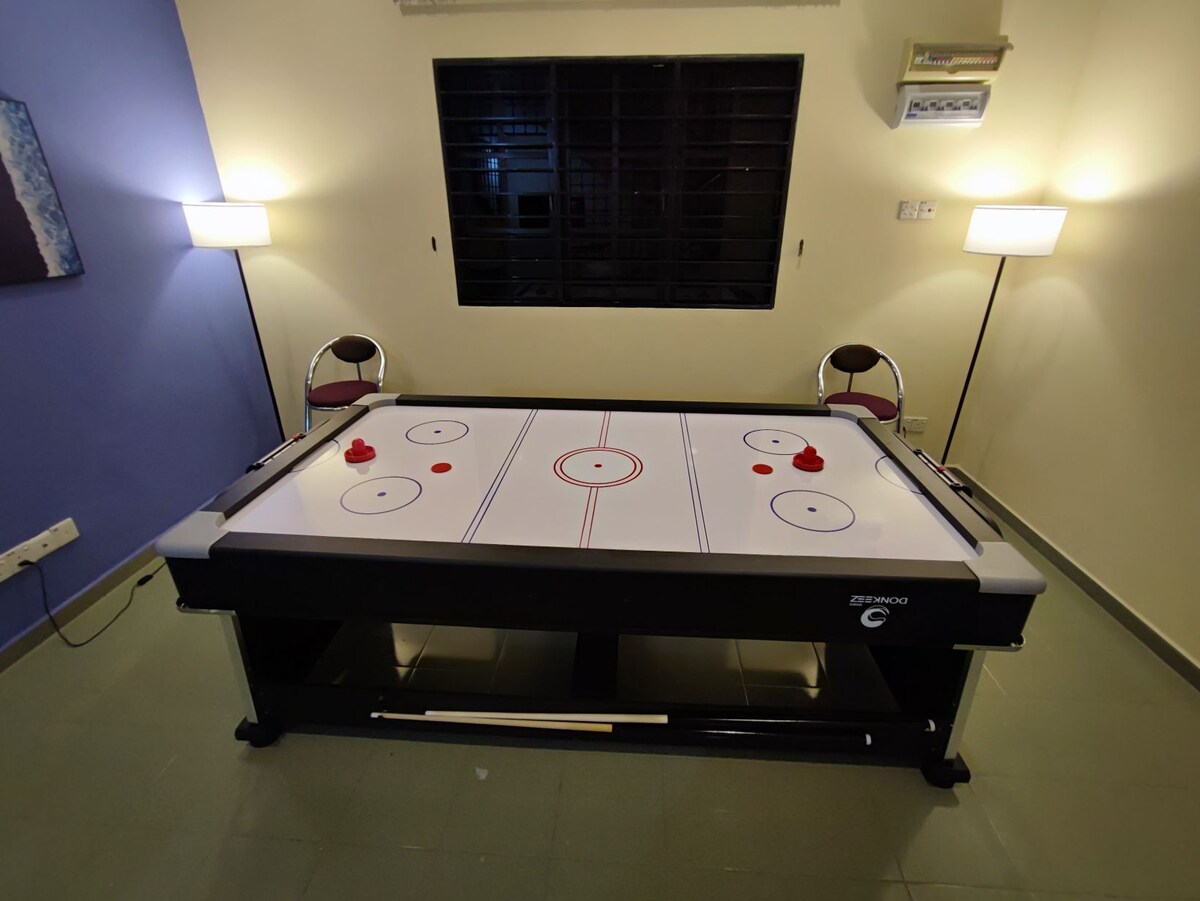 A game room is equipped with a sleek air hockey table, featuring a white playing surface marked with blue lines. Two red pucks are positioned on the table, surrounded by modern chairs and illuminated by soft lighting from lamps on either side.