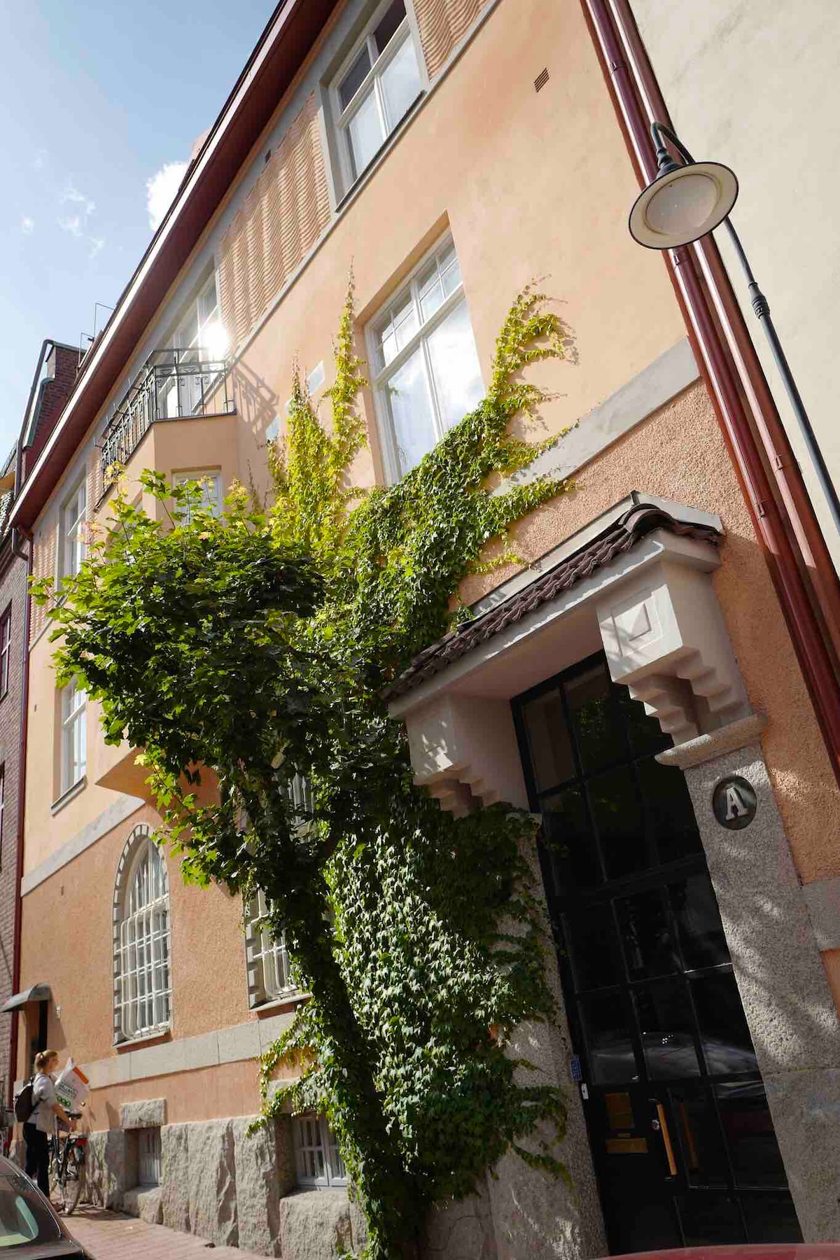 The building features a charming facade with a warm colour palette and decorative architectural elements. Lush green vines climb the exterior wall, adding a touch of nature. Large windows are present, allowing light to fill the interior. A well-maintained entrance is visible at the base.