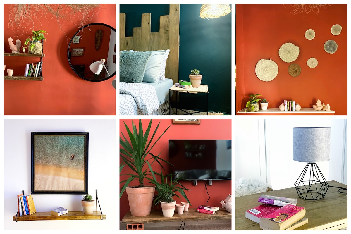 A vibrant living space features a deep orange wall adorned with circular wall decor. A bedside table holds a lamp and plants, while a mirror reflects cozy textures from the bed. A shelf displays small plants and books, adding a homey touch.