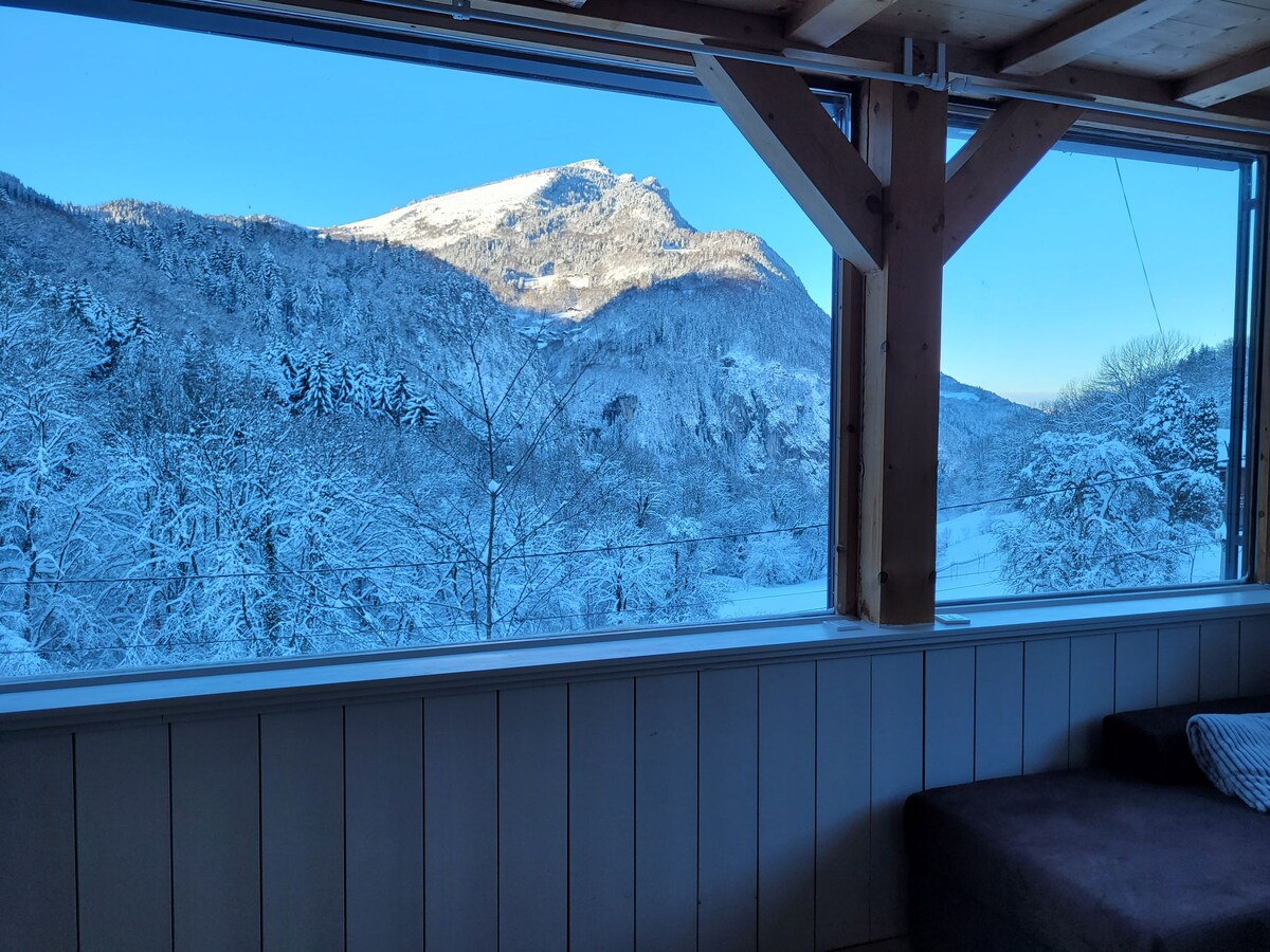A large window frames a stunning winter landscape, showcasing snow-covered mountains and trees under a clear blue sky. The natural light fills the interior space, highlighting the wooden beams that support the structure.