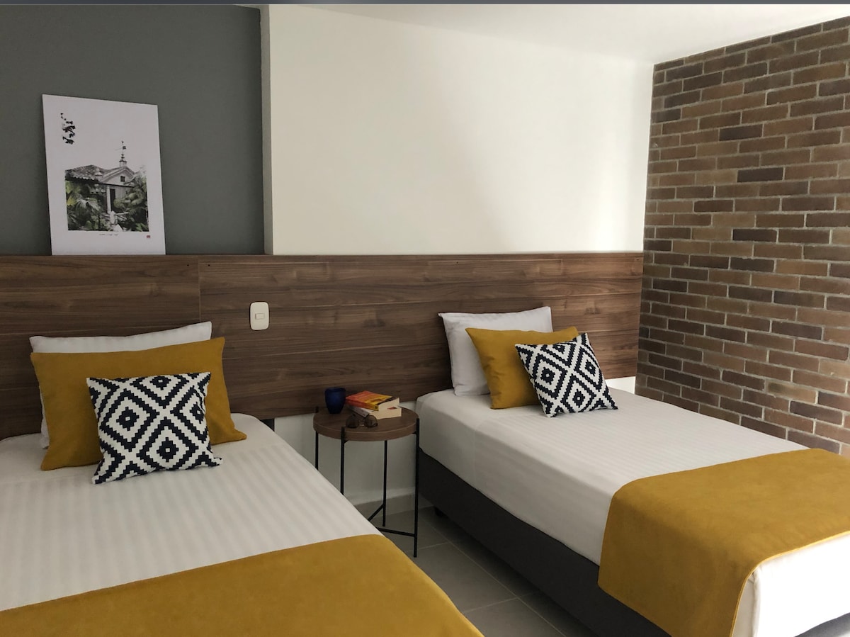 The image displays a well-lit room featuring two beds with white linens and yellow bed throws. A wooden accent wall complements the space, and decorative pillows add a modern touch. A small side table is positioned between the beds, with a few books displayed.