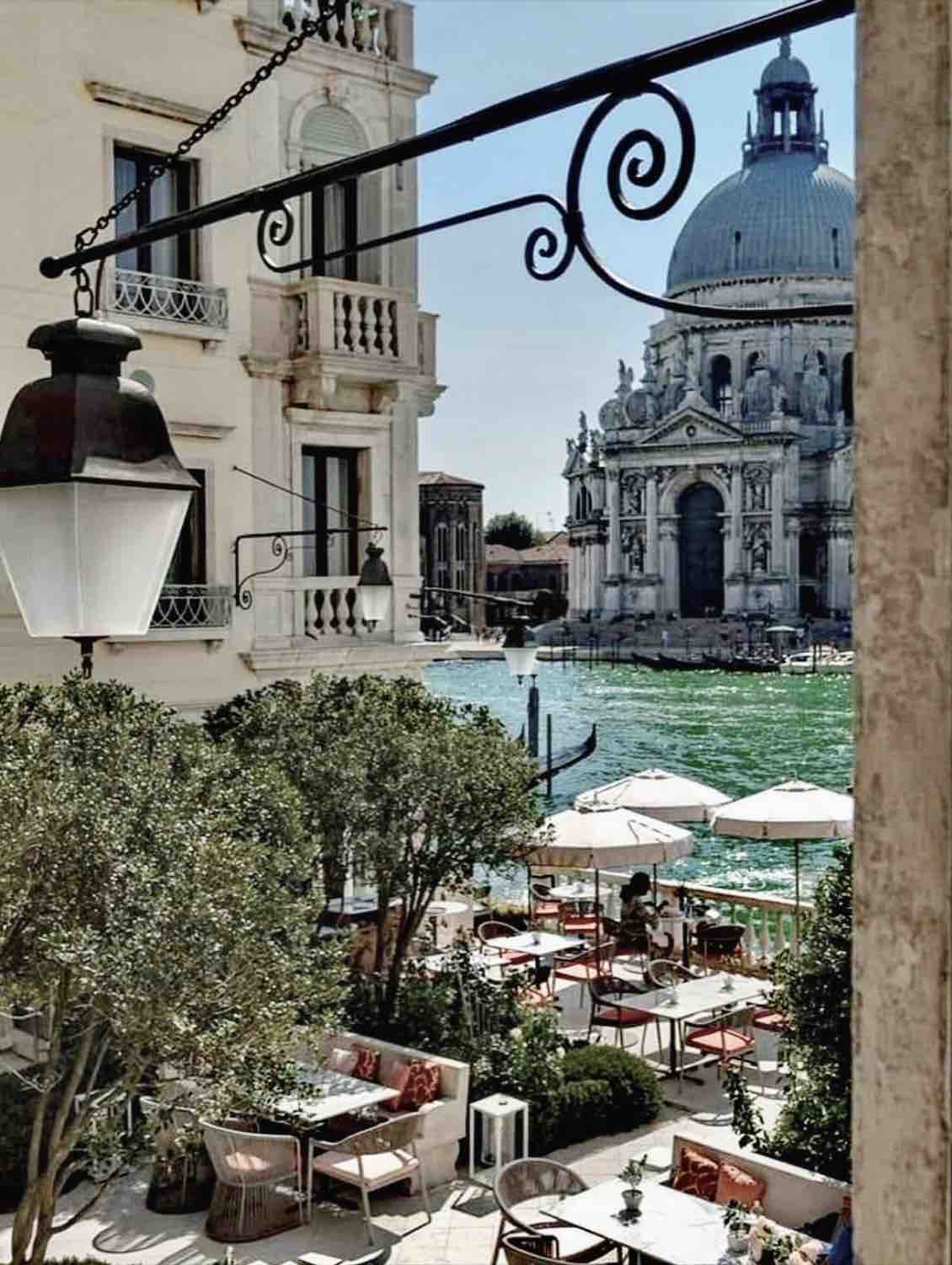 A charming outdoor dining area is visible, featuring tables with umbrellas situated along a canal. The imposing dome of a historic building stands in the background against a bright blue sky, while lush greenery adds a touch of nature to the scene.