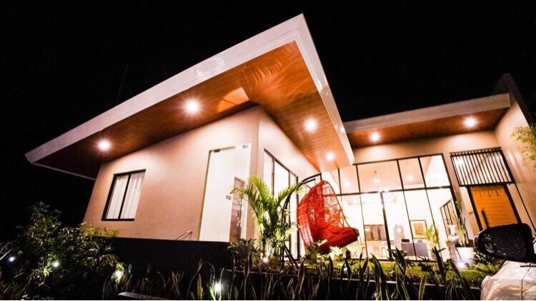A modern house is showcased at night, featuring a distinctive roofline and large glass windows. A vibrant red hanging chair is positioned in the foreground, enhancing the inviting atmosphere. Soft exterior lighting highlights the architectural details and surrounding greenery.