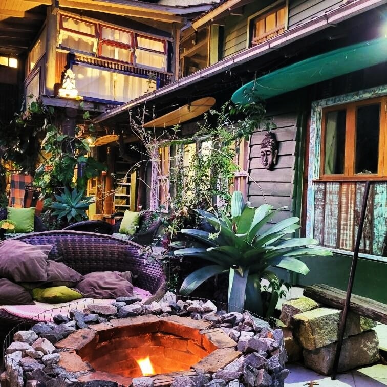 A cozy communal area features a circular stone fire pit at the center, surrounded by comfortable seating and lush greenery. The warm glow from the fire is complemented by the vibrant colors of the surrounding plants and surfboards displayed on the walls.