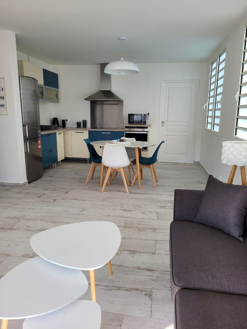 The kitchen and dining area feature modern appliances, including a refrigerator and microwave. A dining table with colorful chairs sits prominently, while a cozy sofa is positioned nearby. Natural light filters through the louvered windows, enhancing the open, inviting space.