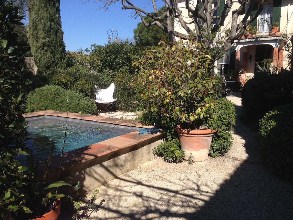 A serene garden features a swimming pool bordered by terracotta pots filled with greenery. A white chair is positioned nearby, surrounded by lush plants. Pathways of gravel lead through the garden, providing a natural and tranquil setting.