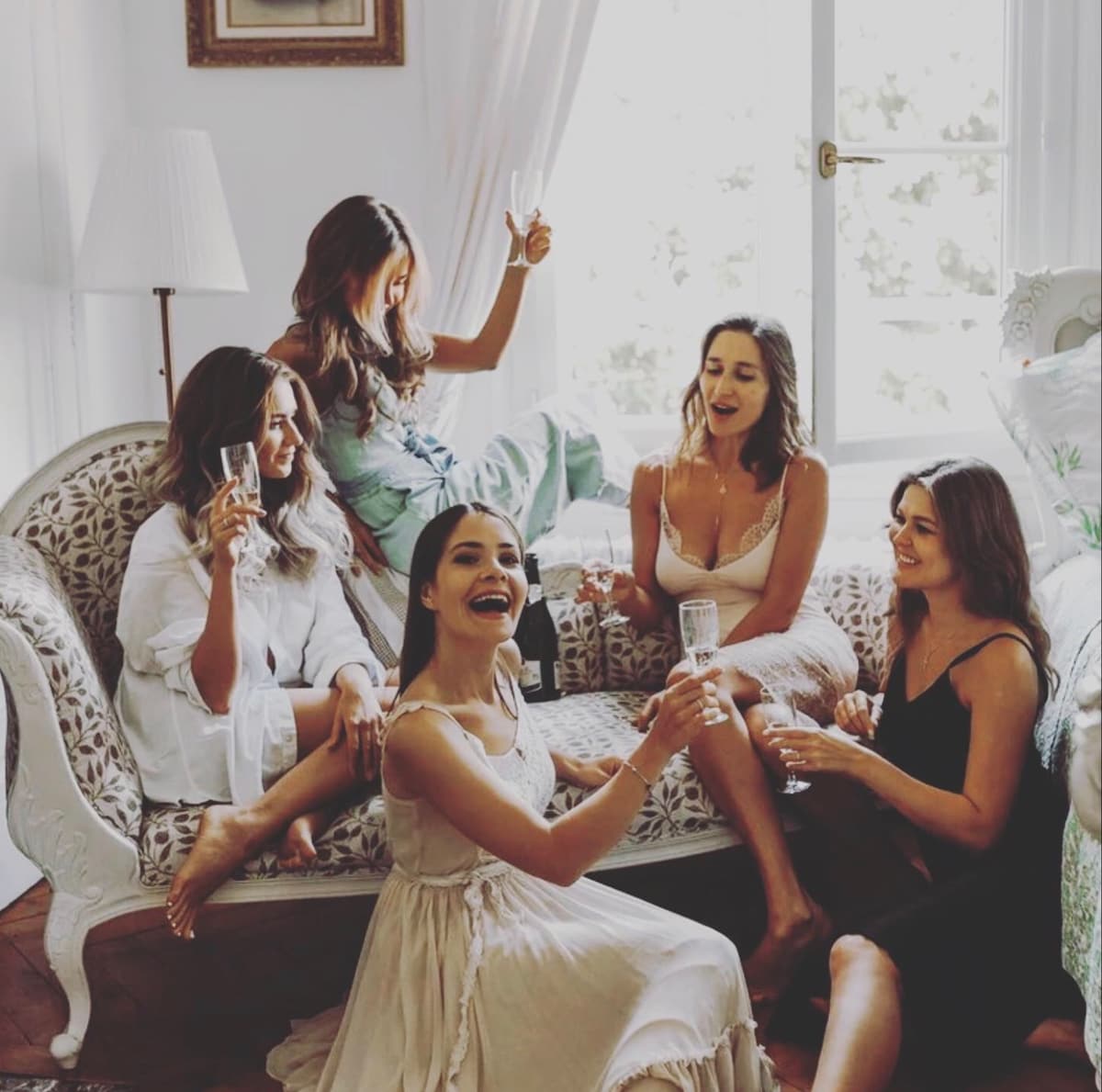 A cozy seating area features a vintage-style sofa with soft upholstery. Five individuals are captured enjoying drinks together, with natural light pouring in through large windows. The atmosphere is relaxed, accentuated by light textiles and a blend of casual attire.