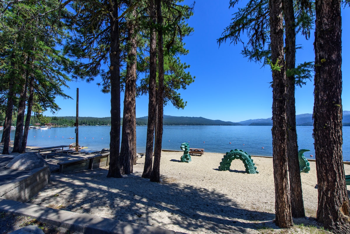 A sandy beach is framed by tall trees, providing a serene setting by the water. The calm lake stretches out towards distant hills, with gentle waves lapping against the shore. Several wooden benches can be seen nearby, offering places to relax and enjoy the view.