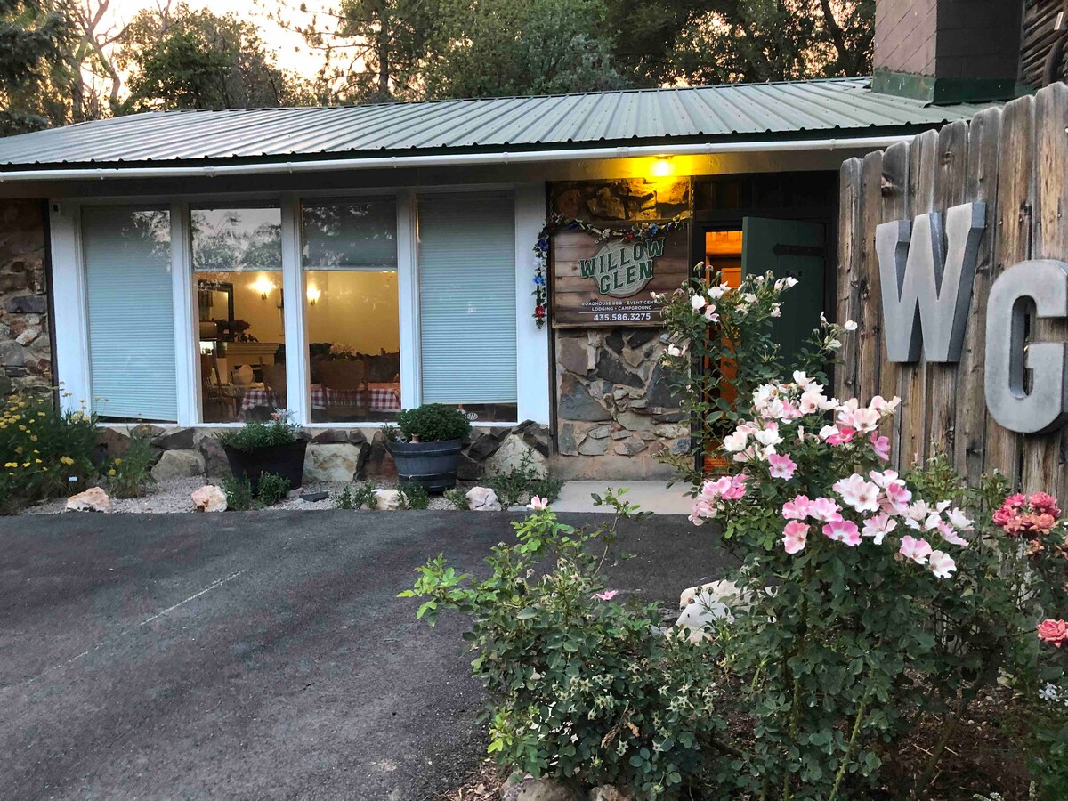 The exterior of the Willow Glen building is presented with a rustic stone façade and a welcoming green metal roof. Colorful flowers line the entrance path, while soft lighting highlights the entrance, inviting guests into a cozy interior setting.