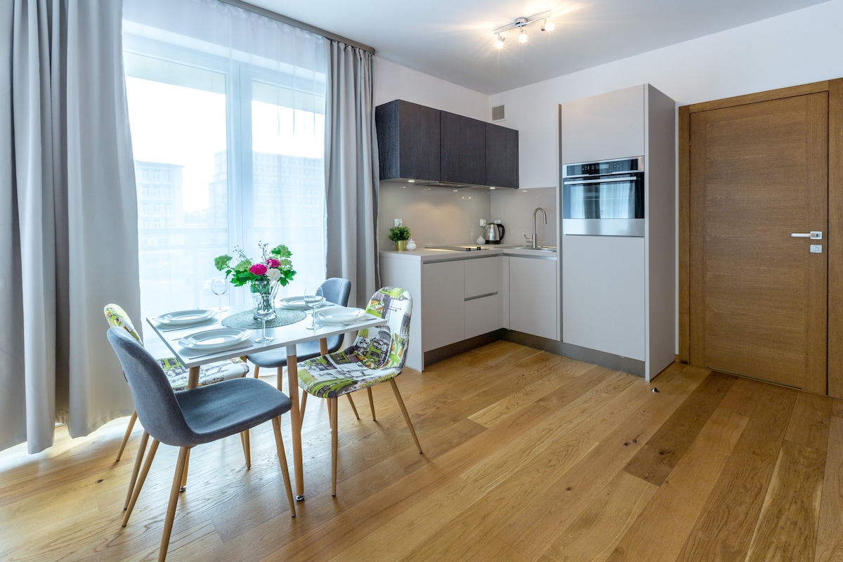 An open-plan kitchen and dining area is displayed, featuring a modern table set for two, with colorful floral accents. Bright light enters through a large window, illuminating sleek cabinetry and stainless steel appliances. Warm wooden flooring adds a cozy touch to the space.