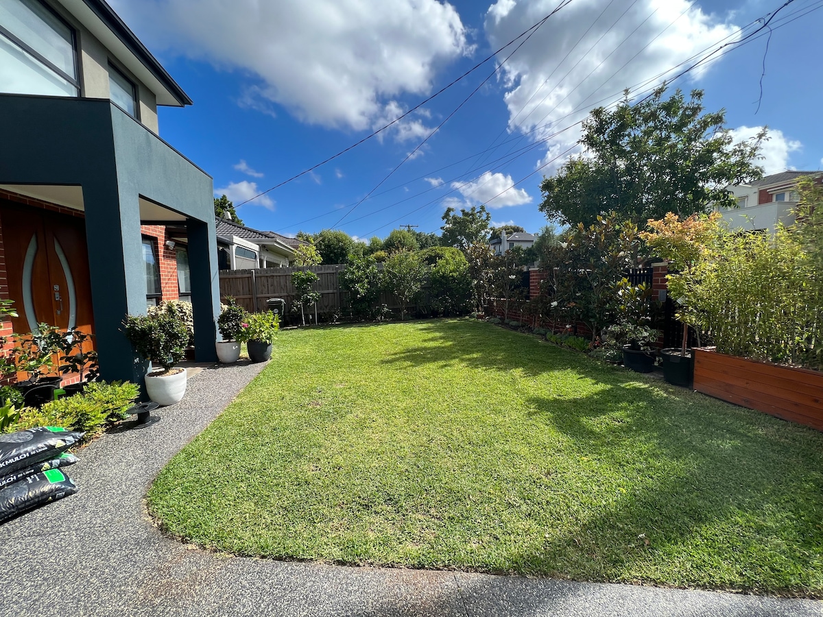 A well-maintained garden area features a lush green lawn bordered by various potted plants. The space is illuminated by natural light, with an expansive blue sky and scattered clouds above. A pathway leads from the house to the garden, promoting access to this outdoor space.