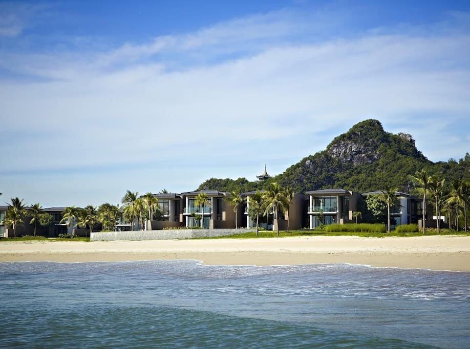 The beachfront villa is showcased against a backdrop of lush greenery and a gentle hillside. Palm trees line the shore, and the waves lapping at the sandy beach create a serene coastal ambiance. The clear blue sky complements the tranquil setting.