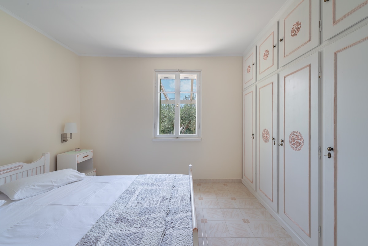 A bright bedroom features a spacious bed with a light quilt. Light enters through a window with four panes, while a decorative wardrobe with pink accents lines one wall. A nightstand and lamp are positioned beside the bed, enhancing the room's functionality.