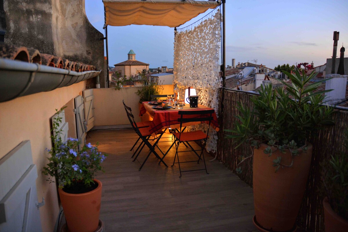 An elevated terrace is featured, adorned with potted plants and a wooden table set for dining. Two folding chairs are positioned around the table, with a soft glow from a lamp. A decorative screen provides privacy while a charming cityscape is visible in the background.