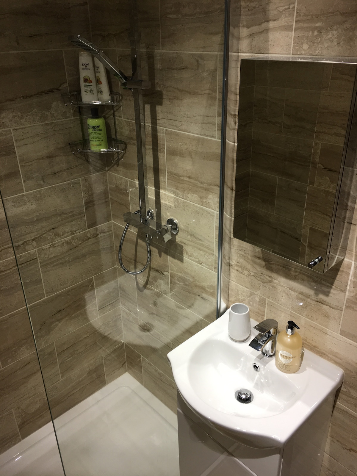 An enclosed en suite bathroom is presented, featuring a rainfall shower with a glass partition. Beige stone tiles line the walls, complemented by a contemporary sink with a sleek faucet. Shelves hold various toiletries, and a mirror reflects the well-lit space.