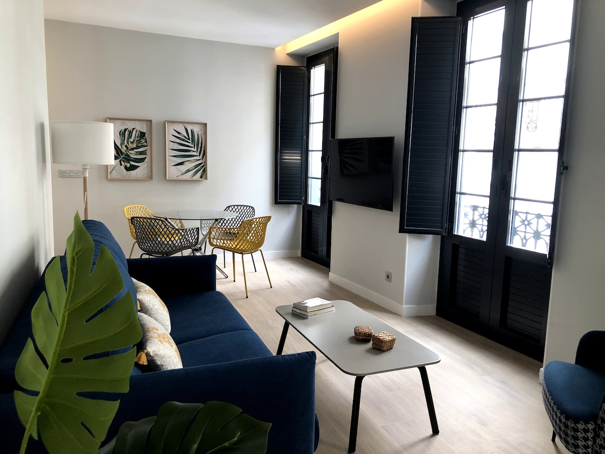 A modern living area is displayed with a blue sofa and a low coffee table. Two yellow chairs are positioned at a dining table, and a wall-mounted television is visible. Tall windows with black shutters allow natural light into the space.