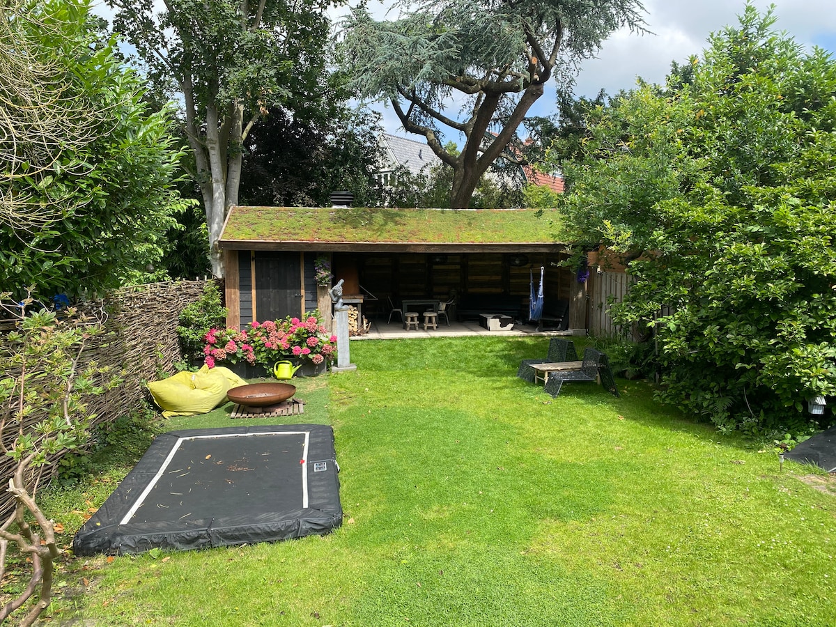 A spacious garden area is seen, featuring a lush green lawn and colorful flowerbeds. A wooden shed sits in the corner, equipped with seating and shade. A trampoline occupies one side, while a cozy lounge chair is positioned nearby for relaxation.