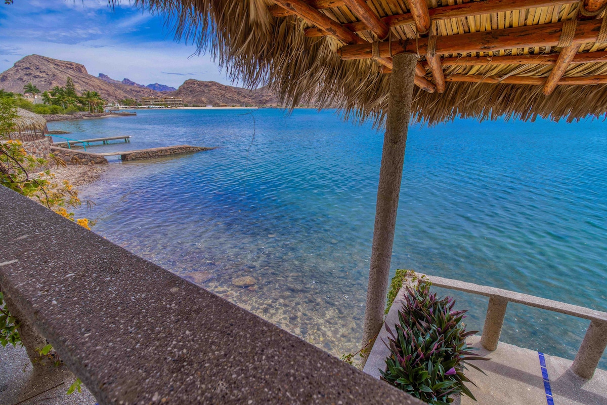 A thatched-roof structure frames a tranquil waterfront view, showcasing clear blue water and rocky shorelines. Lush greenery is visible along the edges, leading to distant mountains under a clear sky. The scene conveys a sense of peaceful natural beauty.