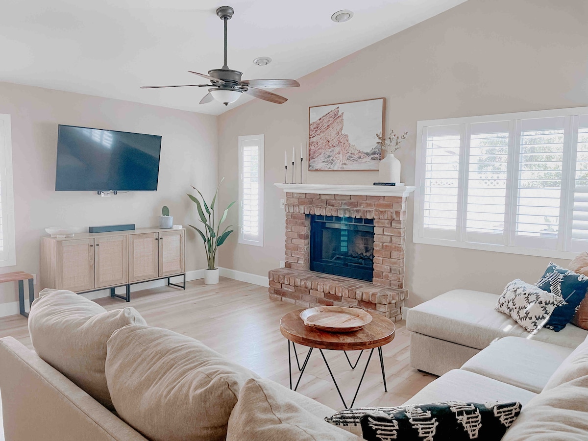 The living area features a sectional sofa and a round coffee table, complemented by a brick fireplace and a wall-mounted television. Light filters through the shuttered windows, adding warmth to the neutral tones of the decor. A green plant is positioned beside the fireplace.