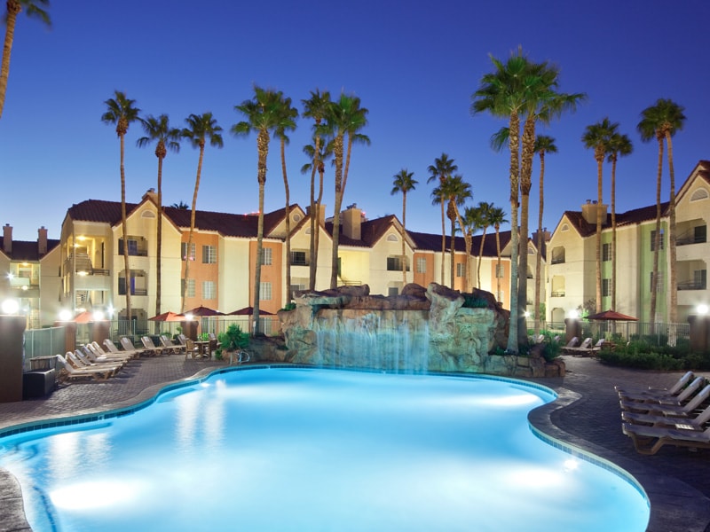 A tranquil swimming pool is illuminated with a soft blue glow, surrounded by palm trees and lounge chairs. A rock waterfall feature adds a natural element, while the resort buildings are softly lit in the background against the evening sky.