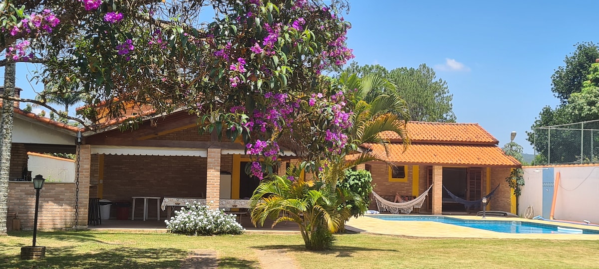 A bright outdoor area is presented, featuring a swimming pool surrounded by a well-maintained lawn. Colorful flowers and greenery enhance the space, while a hammock and shaded seating areas are visible near the house with a terracotta roof.